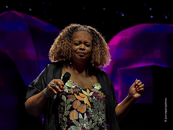 Dianne Reeves hace una pausa mientras canta micrófono en mano y cierra los ojos. Es un plano medio frontal que deja ver el decorado de tonos azules y fucsia.
