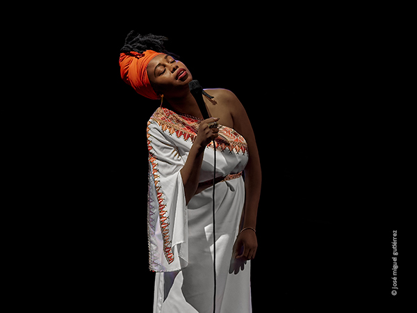 Foto en plano 3/4 en la que vemos a Jazzmeia Horn cantando con la cabeza inclinada hacia su derecha a modo de desmayo. Tiene los ojos cerrados y luce un vestido blanco con forma de túnica y un turbante alargado de color naranja por el que se pueden ver por arriba sus rastas negras.