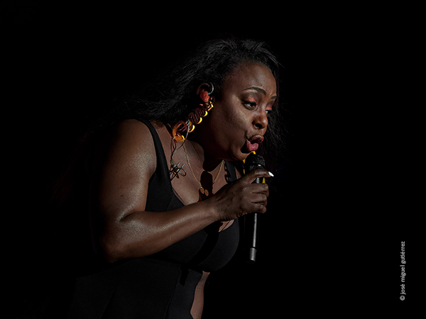 Ledisi canta casi de perfil en la foto de plano medio donde relucen dos enormes pendientes de color amarillo y una de sus uñas de la mano derecha pintadas de un blanco resplandeciente.