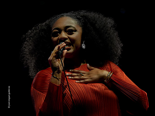 Segundo retrato de Samara Joy del mismo concierto. Plano medio corto en el que se ve cantando micrófono en mano, con su abundante melena negra rizada y con la mano extendida y apoyada en su pecho.