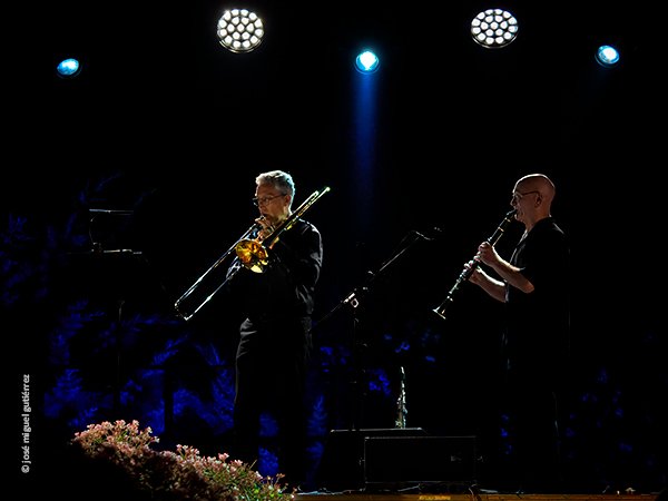 Jan Smets tocando el trombón y a su lado Dirk Descheemaeker tocando el clarinete, ambos de perfil, en una bella imagen con flores, focos y tonalidades azules.