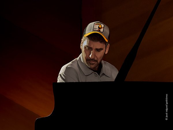 Alex Conde al piano, con gorra que muestra un tucán colorido en su frontal. Retrato que nos muestra su rostro de frente a la cámara en una posición muy atractiva.