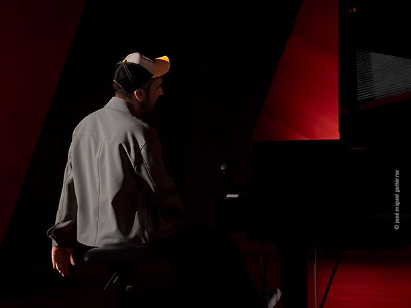 De nuevo Álex Conde al piano en plano largo mostrando su espalda y su brazo izquierdo como desmayado. La iluminación es de intenssos claroscuros con el negro y el rojo intensos y la gorra con su visera amarilla destacando de forma saturada en la imagen,