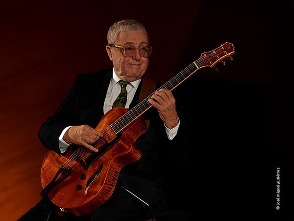 Martin Taylos sentado toca la guitarra y mira hacia su derecha donde se encuentra en fuera de campo Ulf Wakenius. Luce elegancia vistiendo impecable traje negro, corbata y camisa blanca.