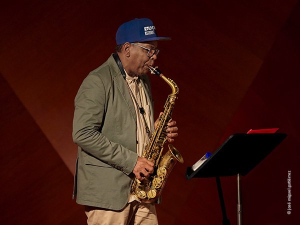 Retrato de Román Filiú tocando el saxo alto de perfil, tocado con una gorra de color azul frente a su atril donde se ve un bote donde puede llevar las láminas de la boquilla del saxo que tiene un tapón azul y rima con la propia gorra.